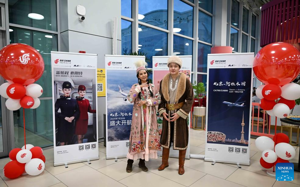 Individu yang mengenakan kostum tradisional Kazakhstan berdiri di hadapan poster laluan udara baharu antara Beijing dan Almaty di Lapangan Terbang Antarabangsa Almaty di Kazakhstan pada 30 Julai 2025. (Xinhua/Li Renzi)
